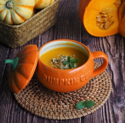 Pumpkin soup in a large orange soup cup that says 'pumpkin cup'. Next to the cup of soup is its lid which looks like the top of a pumpkin. The cup of pumpkin soup is resting on a brown coaster. Behind the cup of soup is a brown basket with a pumpkin inside. Next to the basket is half a pumpkin.